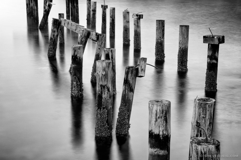 A black and white long exposure photograph of old dock pilings along the Puget Sound at Cummings Park in Tacoma, Washington.