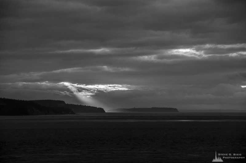 A black and white photograph of sunrays peaking through the clouds over the Puget Sound on a dark Winter day as viewed from Whidbey Island, Washington.