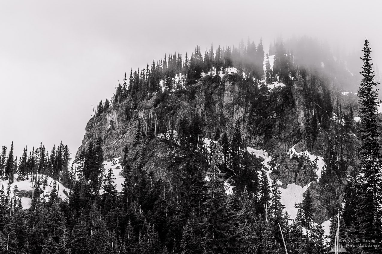 A black and white landscape photograph of a cloud shrouded Crystal Peak as viewed from lower Crystal Lake in the Mount Rainier National Park, Washington.