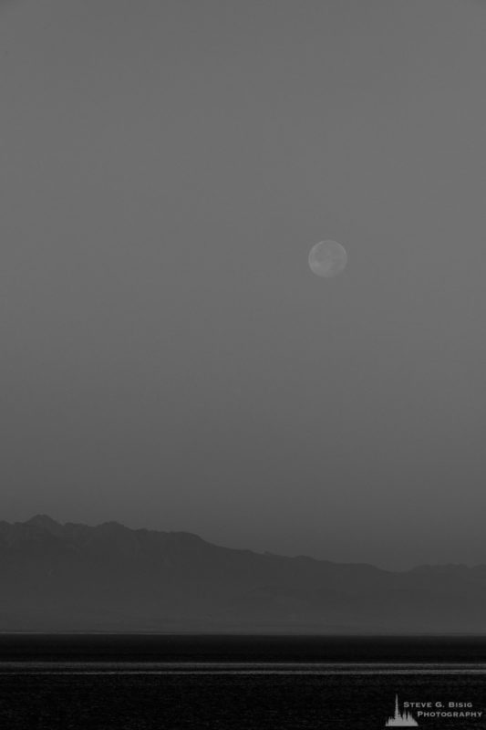 A black and white photograph of the setting full moon on an August morning over Admiralty Inlet as viewed from Ebey's Landing on Whidbey Island near Coupeville, Washington.