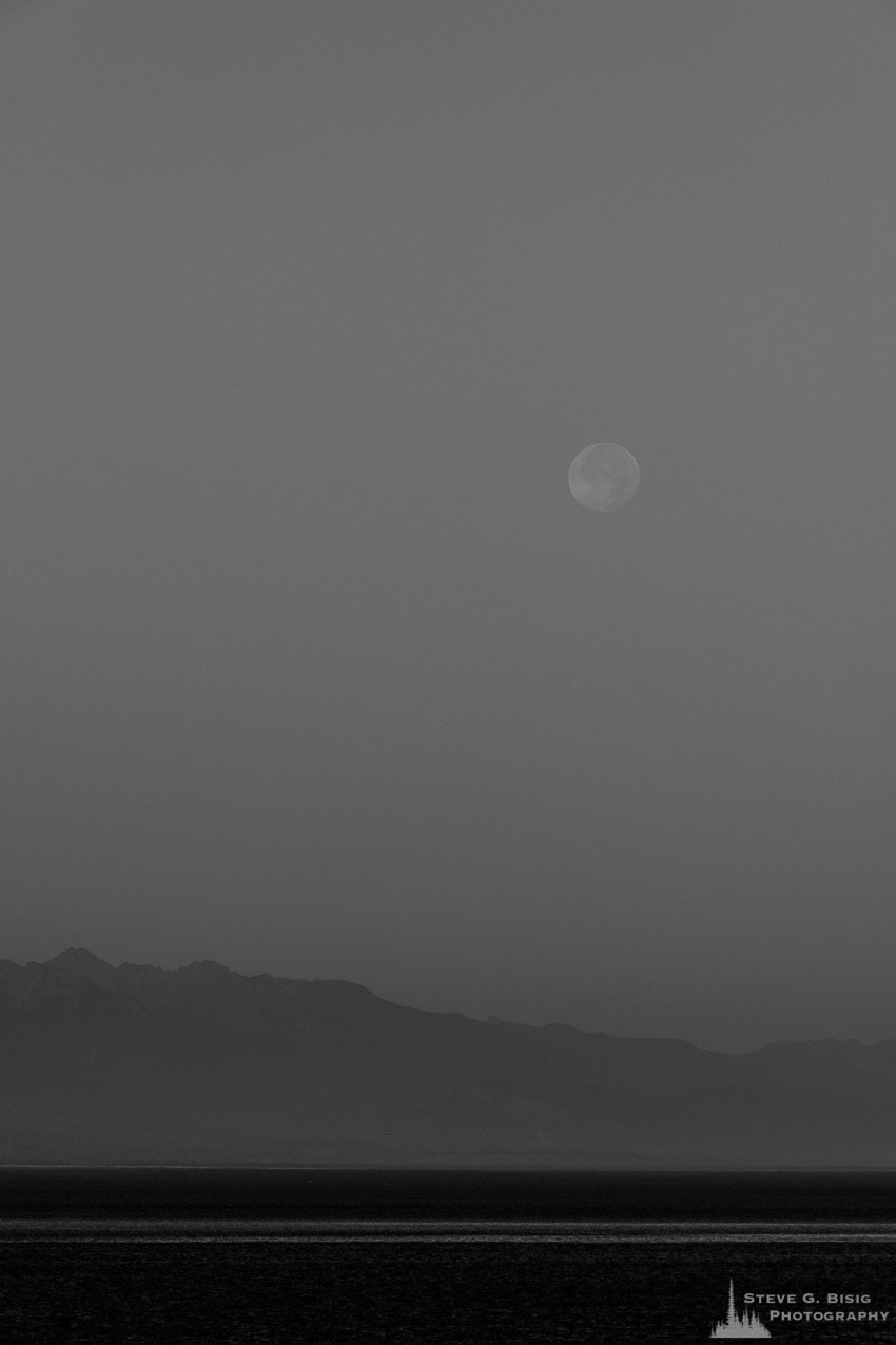 A black and white photograph of the setting full moon on an August morning over Admiralty Inlet as viewed from Ebey's Landing on Whidbey Island near Coupeville, Washington.