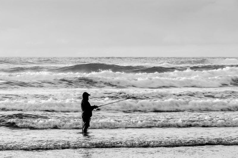 A black and white photograph of a fisherman fishing the Pacific Ocean surf at Tolovana Beach State Recreation Site near Cannon Beach, Oregon.