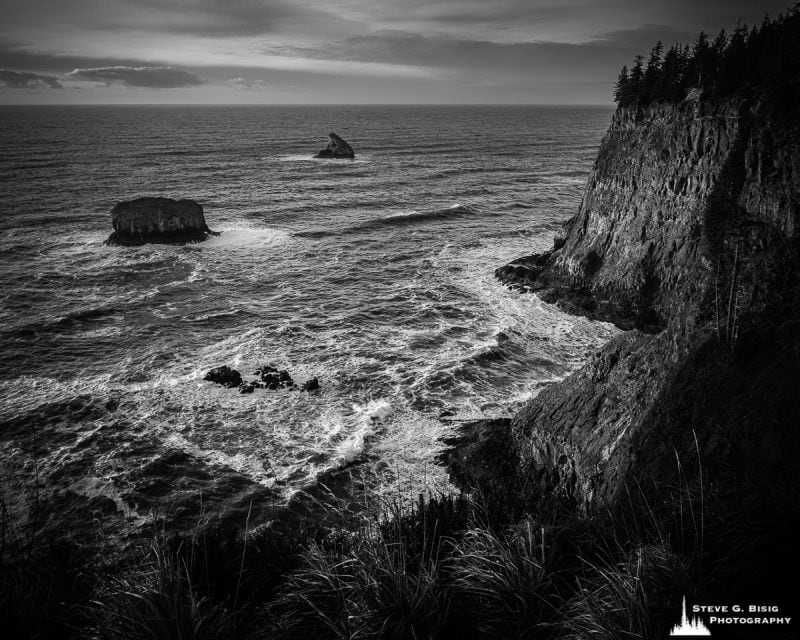 A black and white fine art landscape photograph of the bluffs along the Pacific Ocean coastline and Pillar Rock as viewed from the Cape Meares State Scenic Viewpoint near Tillamook, Oregon.