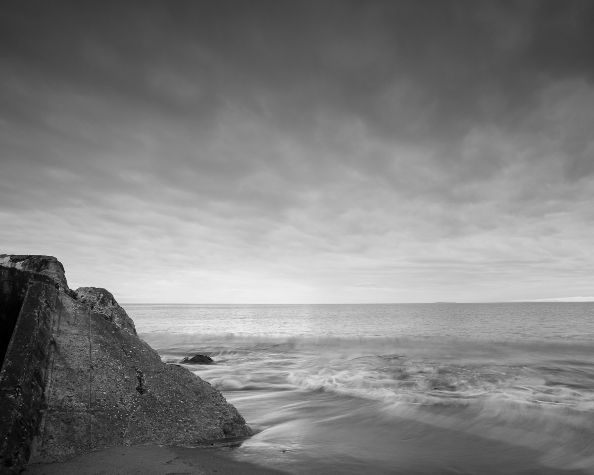 Under the dramatic dance of early autumn clouds, the serene waves of West Beach on Whidbey Island, Washington, gently kiss the rocky shores. The monochromatic tones capture a moment of tranquil solitude, where nature’s raw beauty is both the artist and canvas. Amidst this silent symphony of sea and sky, one can almost hear the whispered secrets of the ancient rocks.