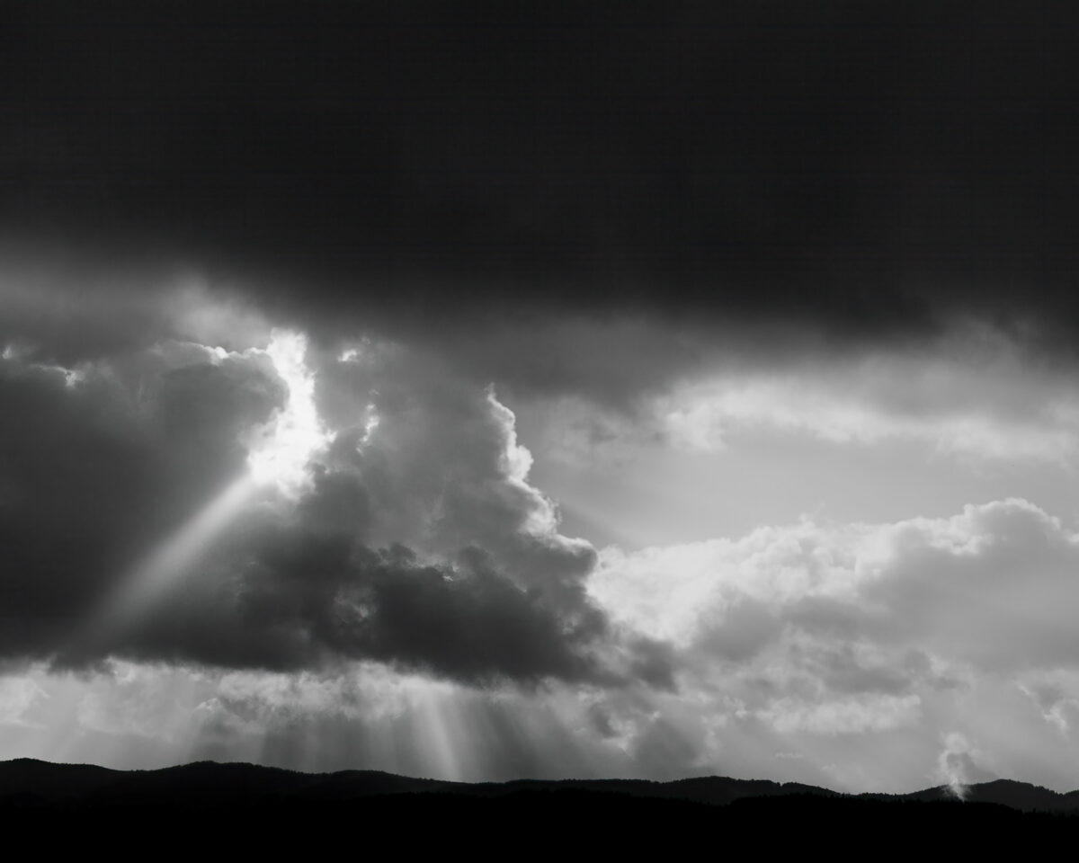 A dramatic scene unfolds over the rugged landscape of Pacific County, Washington. Dark, brooding storm clouds dominate the sky, their ominous presence broken by ethereal shafts of sunlight, contrasting light and shadow. The monochrome palette accentuates the raw power of nature, showcasing the eternal struggle between darkness and light in the ever-changing Pacific Northwest skies.