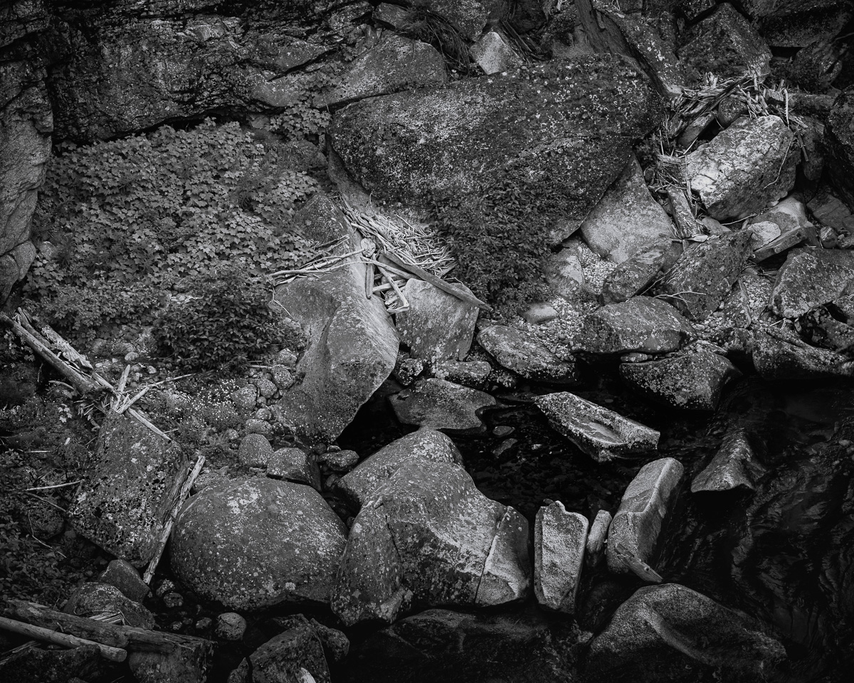 This intimate black-and-white photograph of the Entiat River's rocky bank at Box Canyon reveals a complex tapestry of geological history. Centuries of erosion and natural forces have sculpted this rugged landscape, creating a captivating interplay of textures and shapes. Boulders nestle against jagged rock fragments, while smooth, water-worn stones punctuate the composition. The image invites the viewer to explore the intricate details - from the coarse granularity of ancient rock faces to the delicate patterns of lichen growth. In the shadows, between the stones, a glimpse of the river, a constant presence shaping this ever-evolving natural masterpiece. This scene reminds us of the enduring strength and subtle beauty in even the most rugged corners of our natural world.