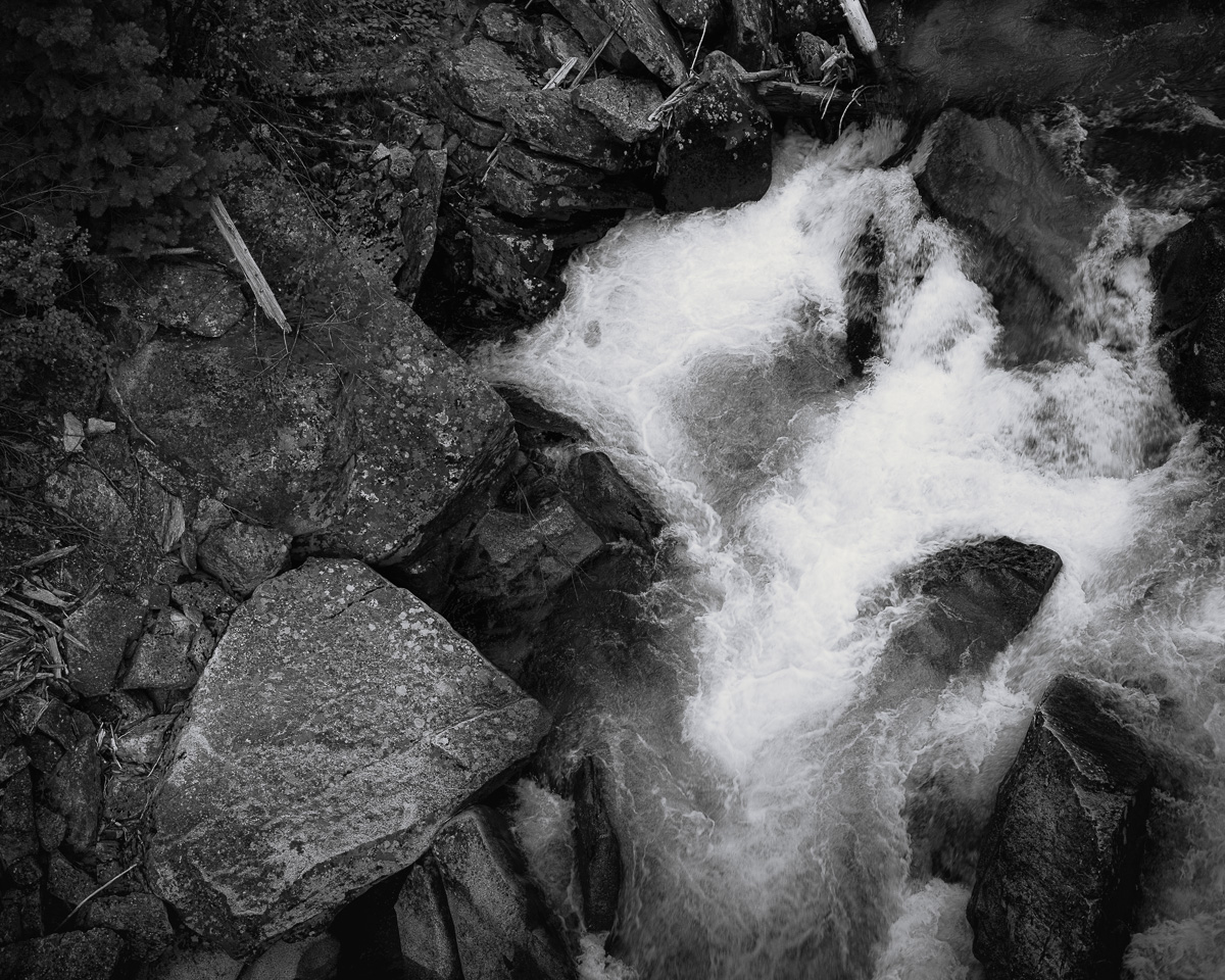 In this black and white photograph, the Entiat River displays its raw power as it crashes through a jumble of ancient boulders. The contrast between the solid, unyielding rock and the fluid, ever-changing water creates a mesmerizing interplay of textures and forms. The exposure transforms the rushing water into a white veil, emphasizing its relentless movement against the dark, immovable stone. This image encapsulates the eternal struggle and harmony between water and rock, showcasing nature's destructive and creative ability to shape the landscape of Box Canyon. The scene serves as a poignant reminder of the constant change and enduring beauty found in wild places.