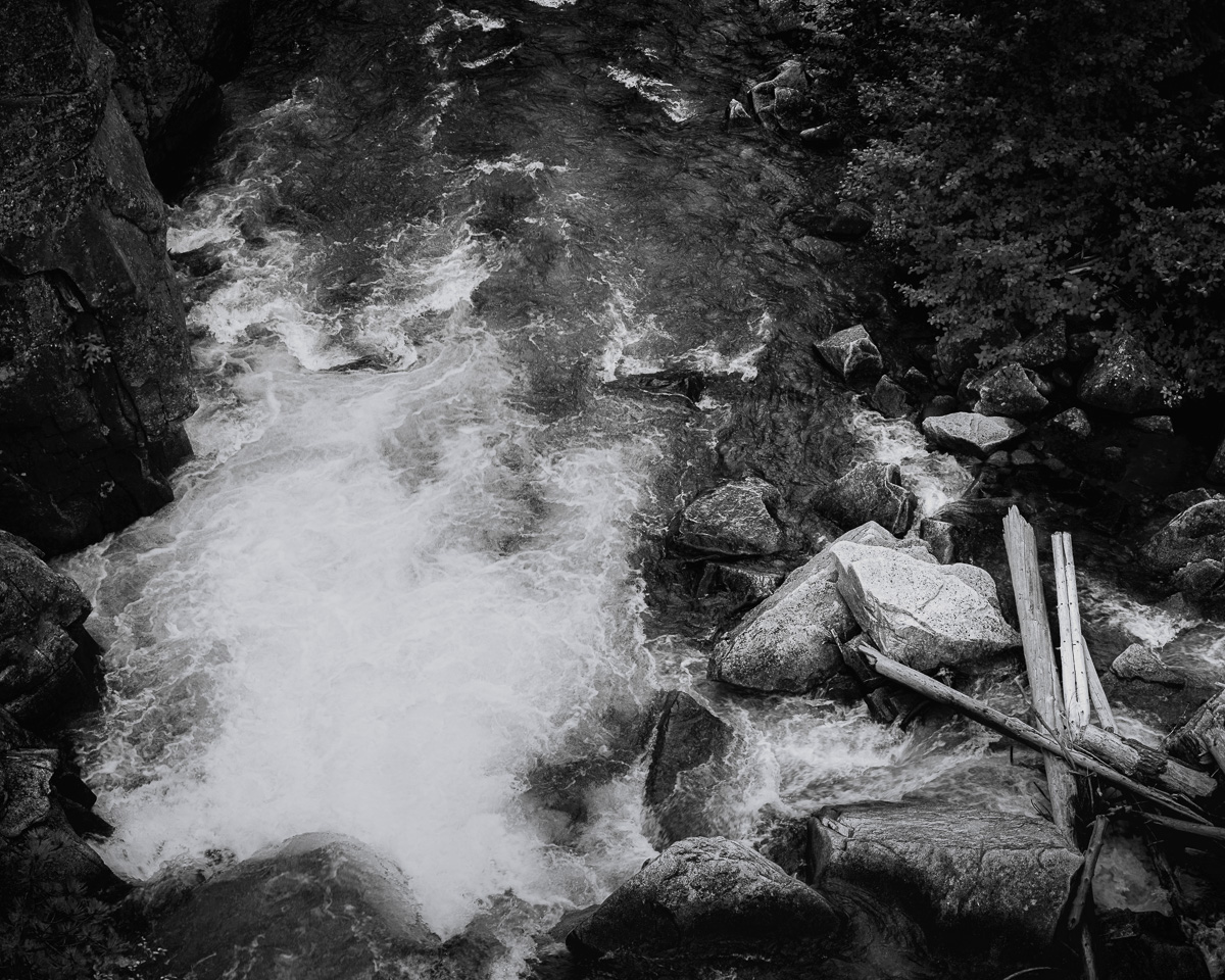 In this black and white photograph, the Entiat River showcases its raw power as it surges through Box Canyon. The exposure transforms the rushing water into a silky, ethereal force, contrasting sharply with the solid, immovable boulders punctuating its path. Rocks line the riverbank, silently witnessing eons of erosion and the river's persistent sculpting of the landscape. The interplay of light and shadow emphasizes the water's tumultuous journey, creating a sense of perpetual motion and timeless struggle between liquid and stone. This image encapsulates the wild spirit of the Entiat River, inviting viewers to contemplate the ceaseless energy and beauty of untamed nature in the heart of Washington's rugged terrain.