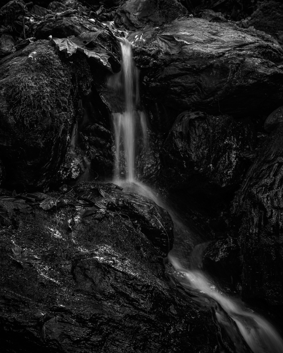 A black-and-white photograph of a small seasonal creek quietly flowing over weathered rocks. The gentle movement of the water contrasts with the dark, textured stones. This tranquil scene captures a fleeting moment and offers a peaceful escape into the wilderness near Darrington, Washington.