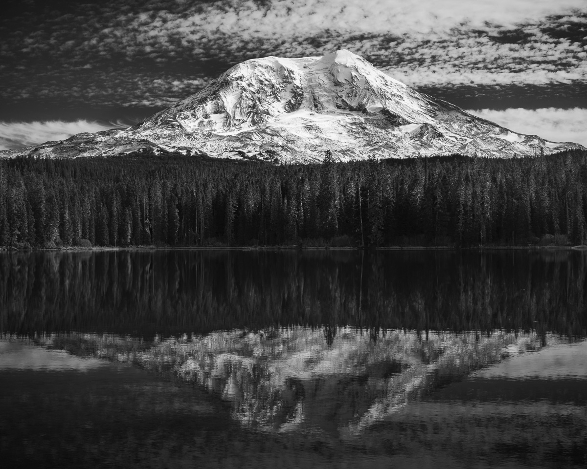 Fresh snow blankets Mt. Adams, reflecting on Takhlakh Lake, Washington. The still water doubles the mountain's presence, capturing the peak's grandeur and the forest's delicate textures. This is autumn's quiet arrival, painted in shades of light and shadow.