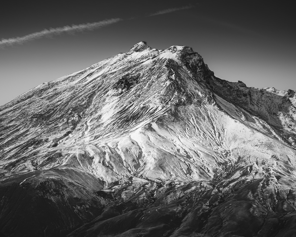 A fresh dusting of autumn snow reveals the rugged textures of the East Crater Rim of Mount St. Helens, etched sharply against the sky. The black and white tones emphasize the raw beauty of the volcanic landscape, showcasing every ridge and shadow. Captured in a quiet moment, the mountain stands both majestic and haunting, its silence echoing tranquility, a silent testament to nature's power and resilience.