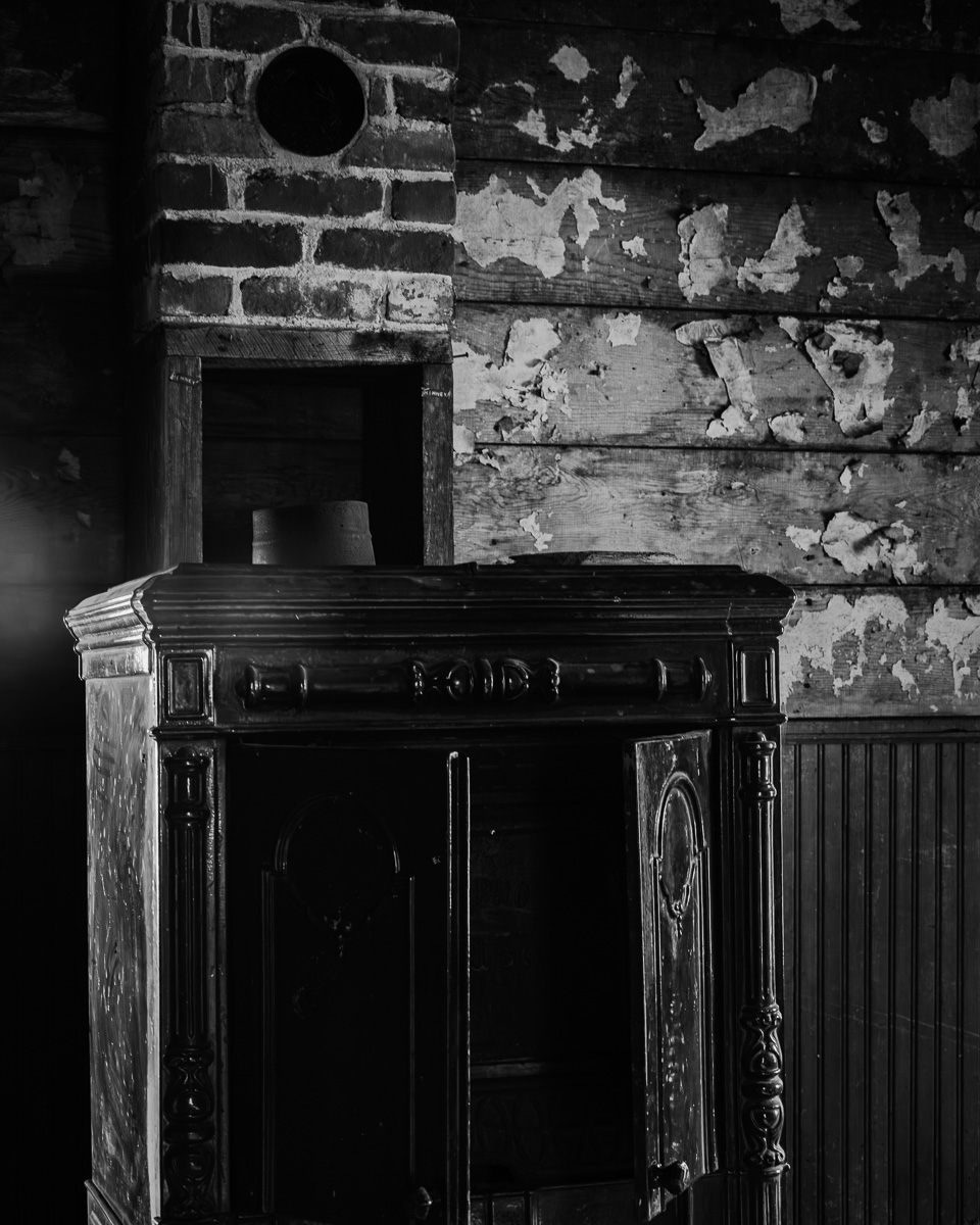 This image invites you into the Whitcomb-Cole Log House in Washington, where a vintage wood stove nestled in the corner resonates with the resilience and resourcefulness of early settlers. The weathered textures and shadows evoke a sense of warmth and history, creating a captivating piece that adds rustic charm to any collection.