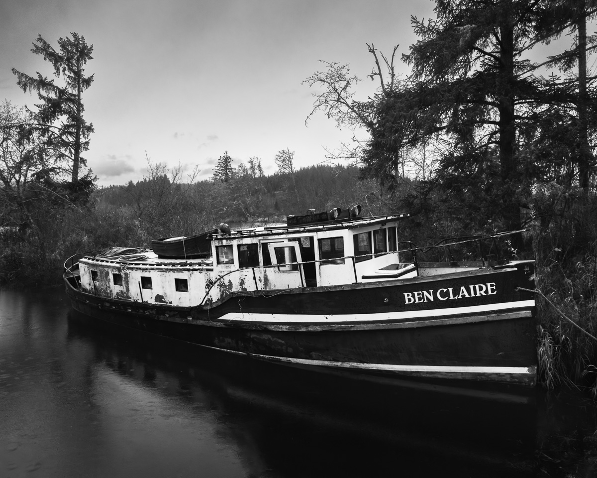 "Moored in Silence" captures a moment of tranquility along the Deep River. The vessel, "Ben Claire," rests still among the trees, inviting reflection on journeys past and the beauty of solitude. This black-and-white photograph layers its vintage allure with a hint of maritime nostalgia, bringing a serene energy to any space.