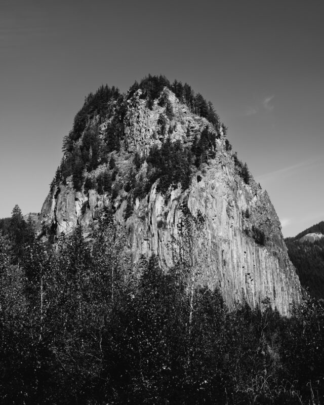 Black and white photograph of Beacon Rock, Washington, showcasing the rocky formation with trees in the foreground, conveying nature's resilience.