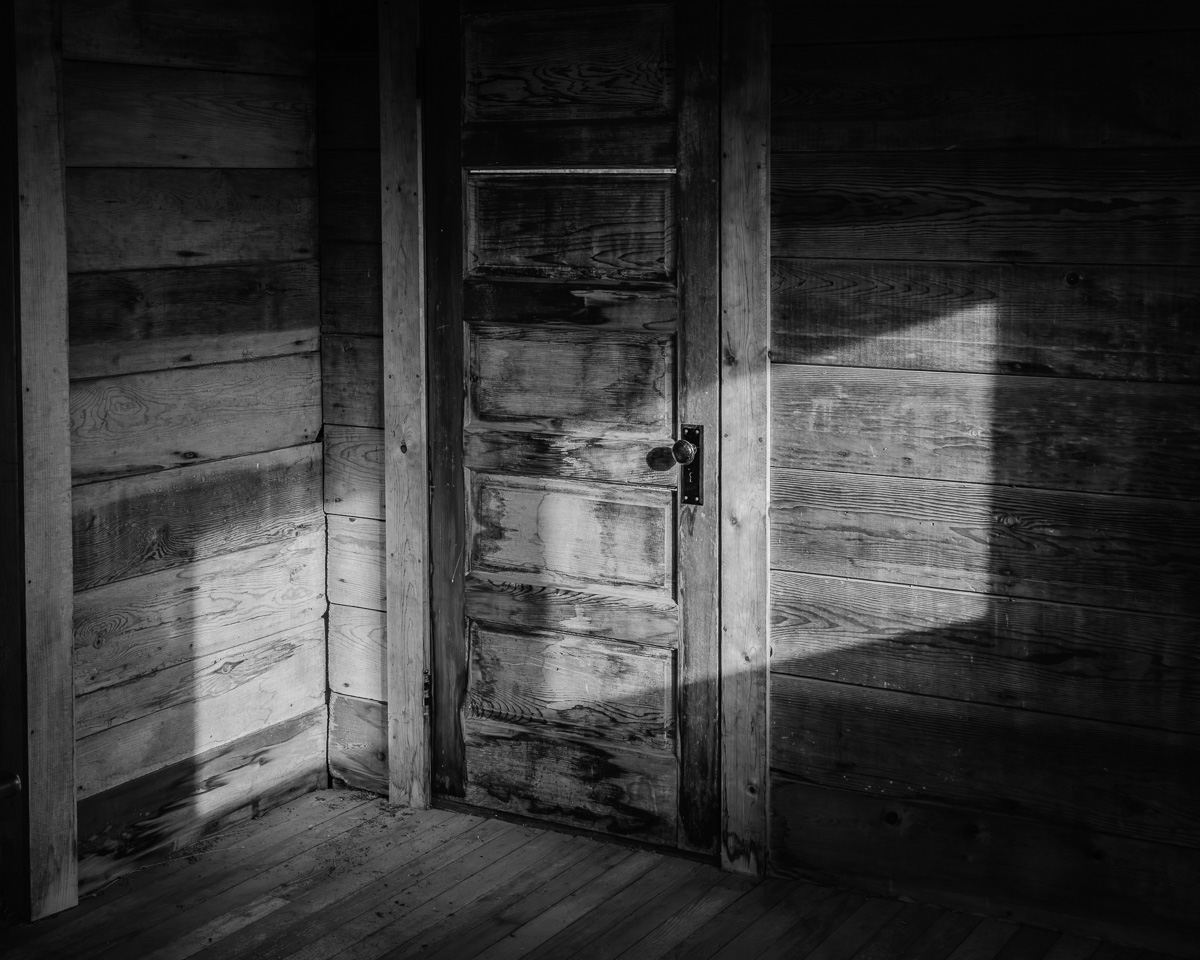 This image invites a moment of quiet reflection, with its shadowy interplay cast against a rustic wooden door. The contrast evokes a lingering sense of mystery, hinting at the stories held within the walls of the Whitcomb-Cole Log House. It's a thoughtful addition to any space seeking tranquility and connection to the past.