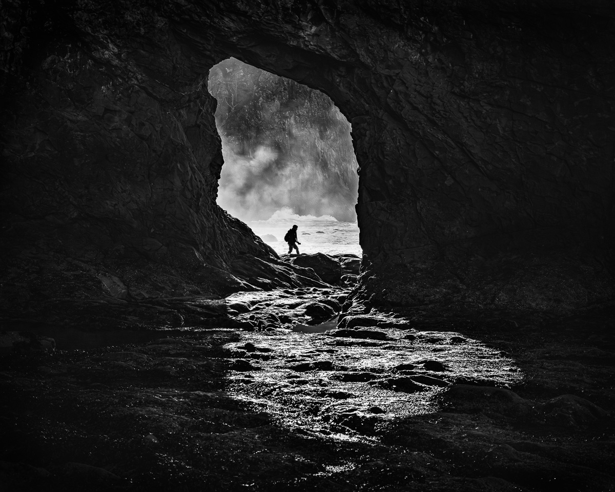 Discover the mystery and allure of Rialto Beach in this captivating black-and-white photograph. A lone figure passes beyond the “hole in the wall,” framed by the rugged contours of a natural cave, embodying the serene beauty of Washington's winter coastline.