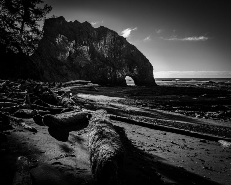 In this black-and-white photograph, the winter coastline of Rialto Beach in Olympic National Park, Washington, is revealed as shadows dance across the sand and rocks. The dramatic arch stands as a testament to nature's artistry, inviting reflection and tranquility.