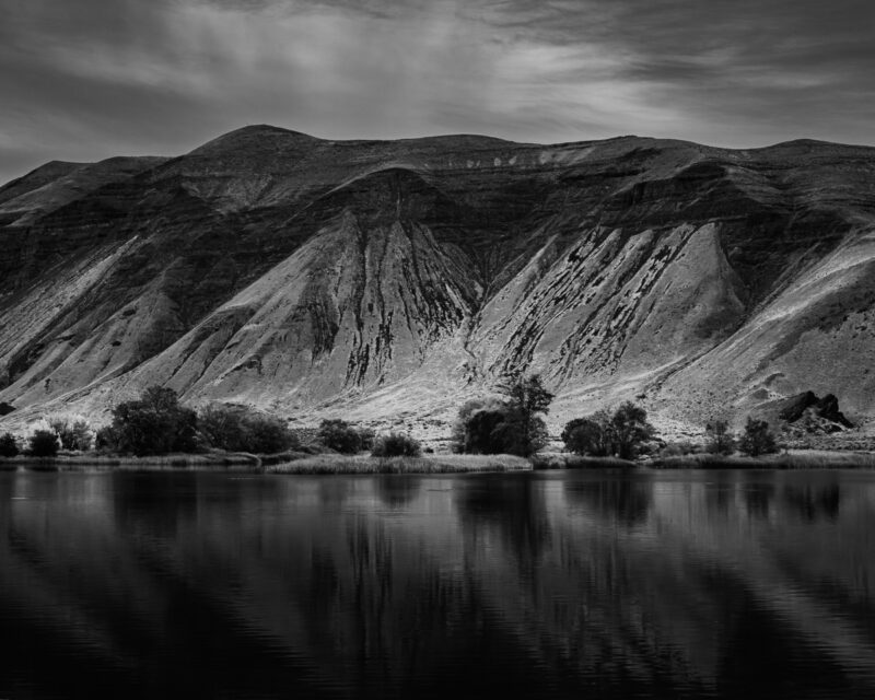 This black-and-white landscape photograph captures the serene beauty of Burkett Lake, cradled beneath the Saddle Mountains in Lower Crab Creek Coulee, Washington. Reflecting rugged slopes and soft ripples, it evokes a quiet balance between nature’s enduring strength and gentle stillness—a mindful connection to the land.