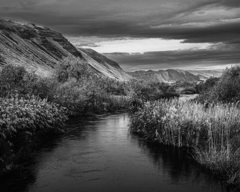 This black-and-white photograph captures the flowing stillness of Crab Creek in Washington's Lower Crab Creek Coulee. Framed by windswept ridges and autumn vegetation, the scene embodies solitude—inviting quiet reflection through nature's gentle forms.