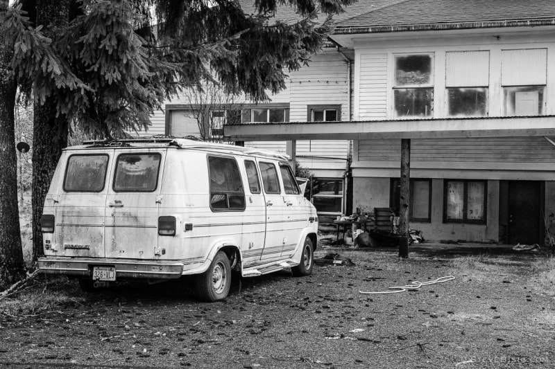 A black and white photograph of an abandoned Cheverolet van at the now abandoned Dryad School in the small Lewis County community of Dryad, Washington. At some point in the recent past, the school was converted into residential living quarters (maybe a four-plex), but has since became vacant (or so it appears).