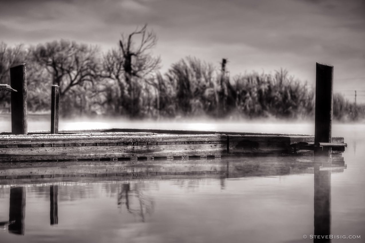 A black and white photograph of a fishing dock reflecting into the water in Ellensburg, Washington. Mist rising over the water.