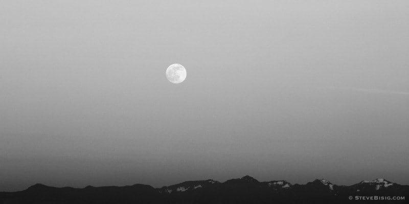 A black and white photograph of the May 2014 full moon rising over the Washington State Cascade Mountains as viewed from Tacoma, Washington.