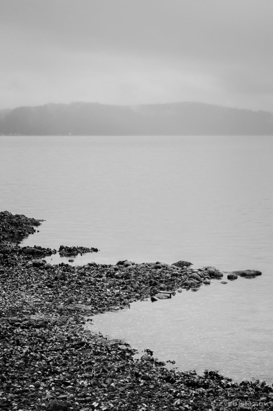 A black and white photograph of the rocky beach along Hood Canal at the Triton Cove State Park near Brinnon, Washington. The Kitsap Peninsula in the distances.