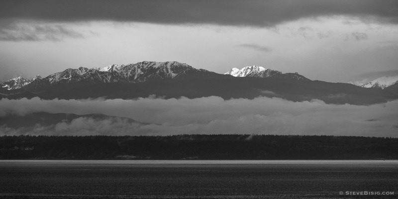 A black and white photograph of the morning sun shining on the Olympic Mountains on a cloudy Spring day. Viewed from across the Puget Sound at Fort Casey State Park on Whidbey Island, Washington.